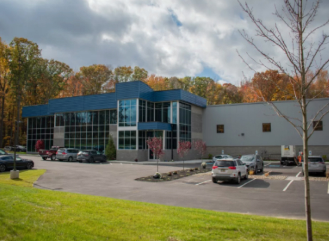 A modern commercial building with a blue roof and a large glass facade is shown on a cloudy day. A parking lot in front of the building contains several parked cars.